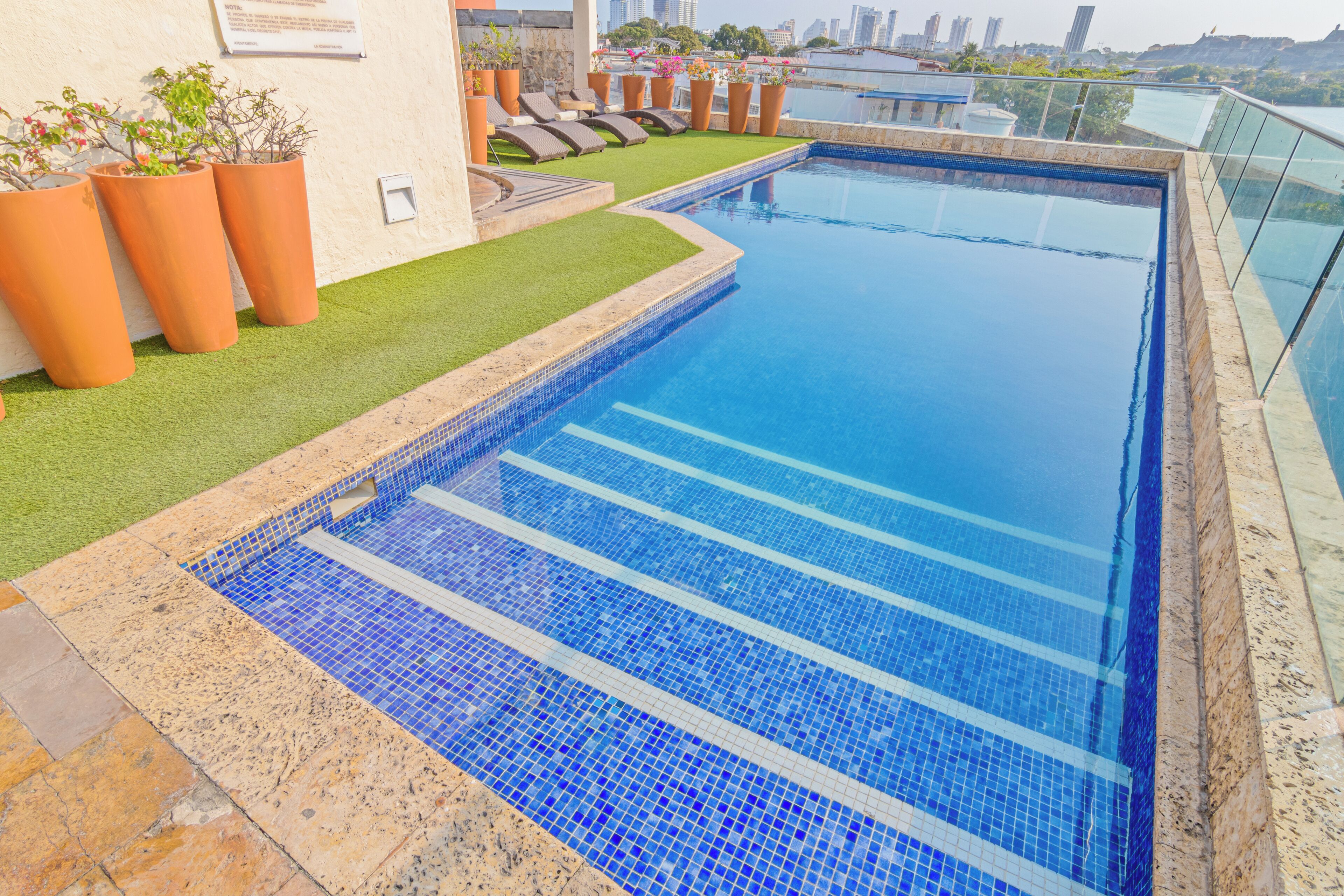 A rooftop pool with clear blue water, surrounded by terracotta planters and a grassy area with lounge chairs.