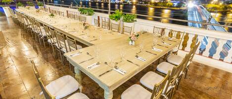 Una larga mesa de comedor con sillas, preparada para una comida, con vista a un río y luces de la ciudad al fondo.