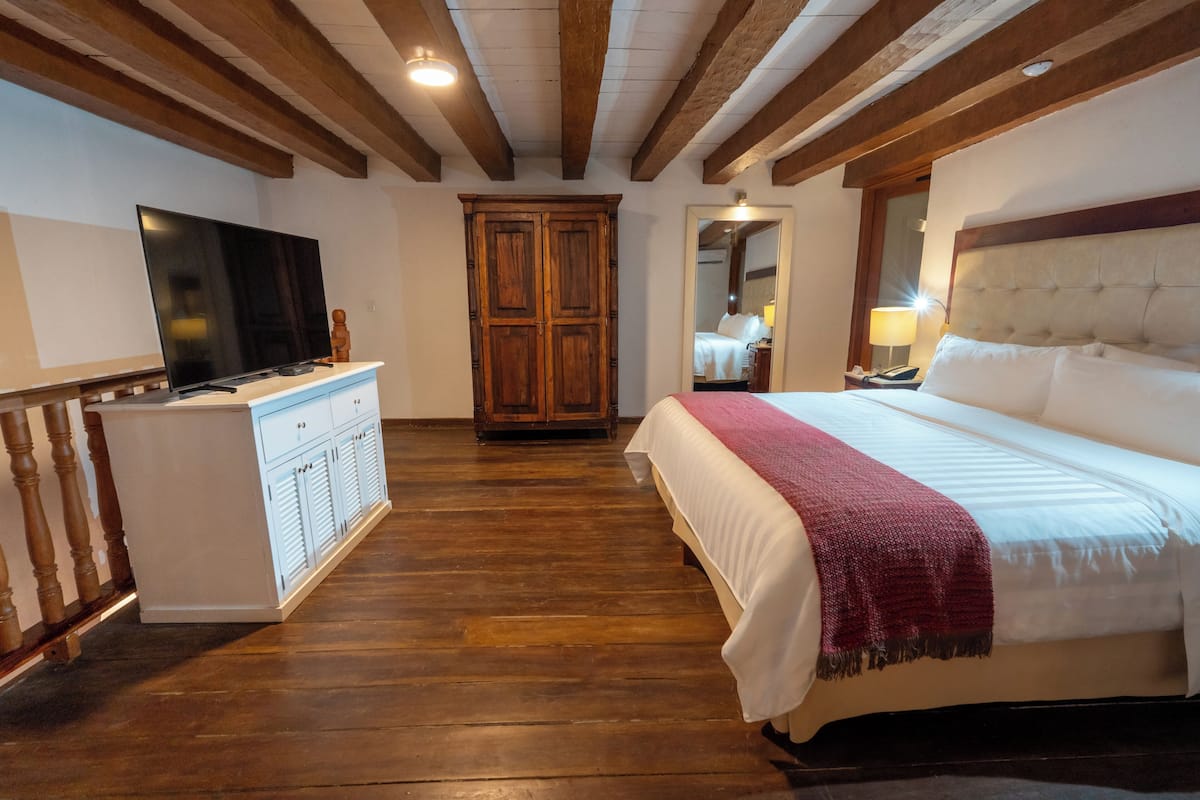 A hotel room with a wooden floor, a bed with white bedding and a red throw, a TV on a white cabinet, and a wooden wardrobe.