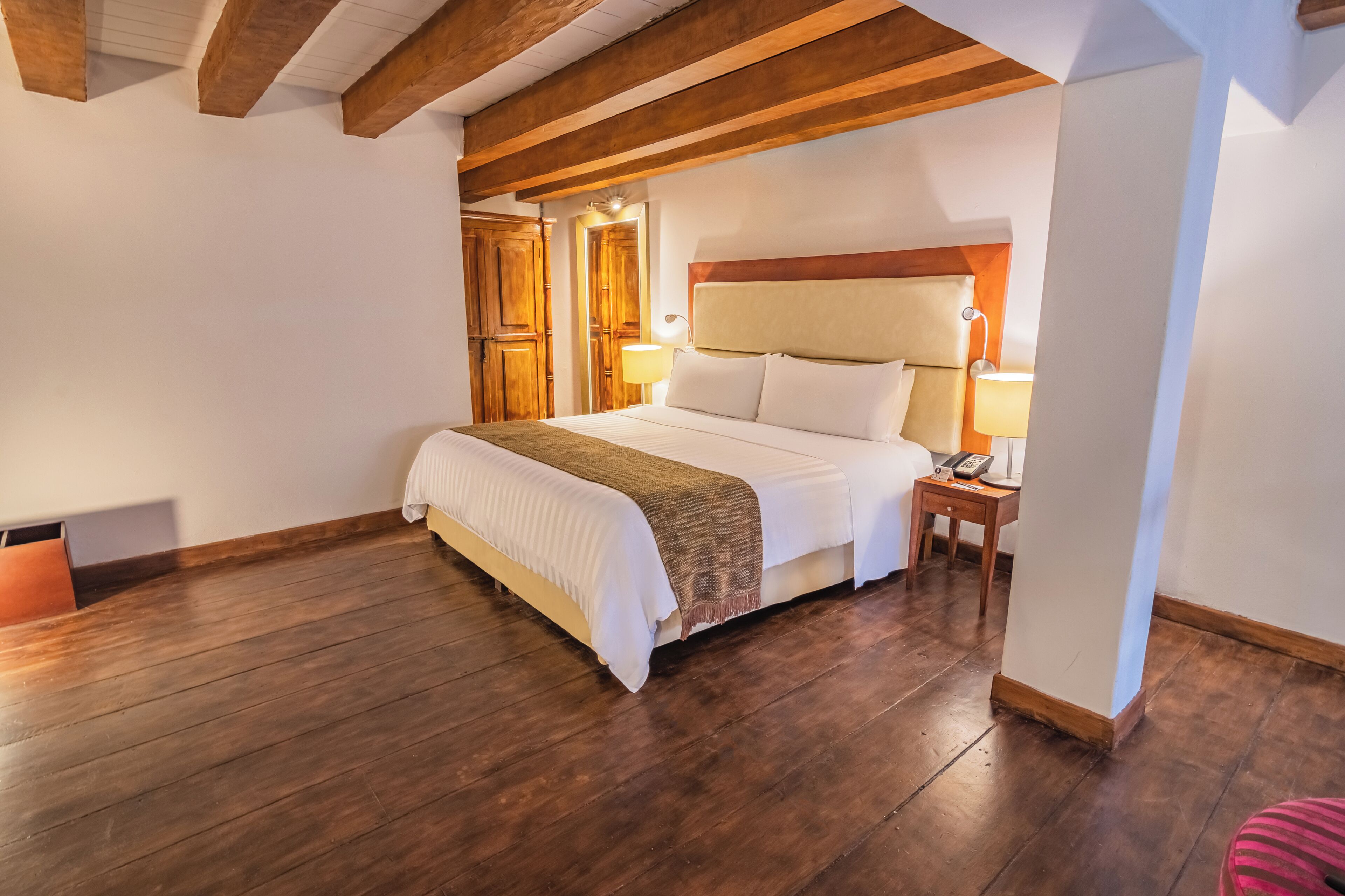 A bedroom with a large bed, wooden beams, a nightstand with a lamp, and a wooden door.