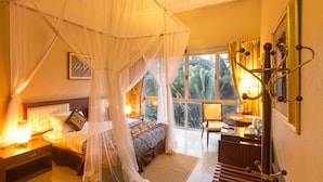Une chambre à coucher avec un lit à baldaquin, un bureau avec une lampe et une vue sur la végétation tropicale, visible à travers de grandes fenêtres.