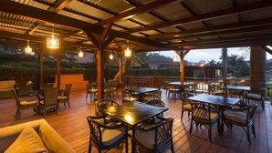 Une terrasse extérieure avec des tables et des chaises en bois, un espace salon couvert et des guirlandes lumineuses.