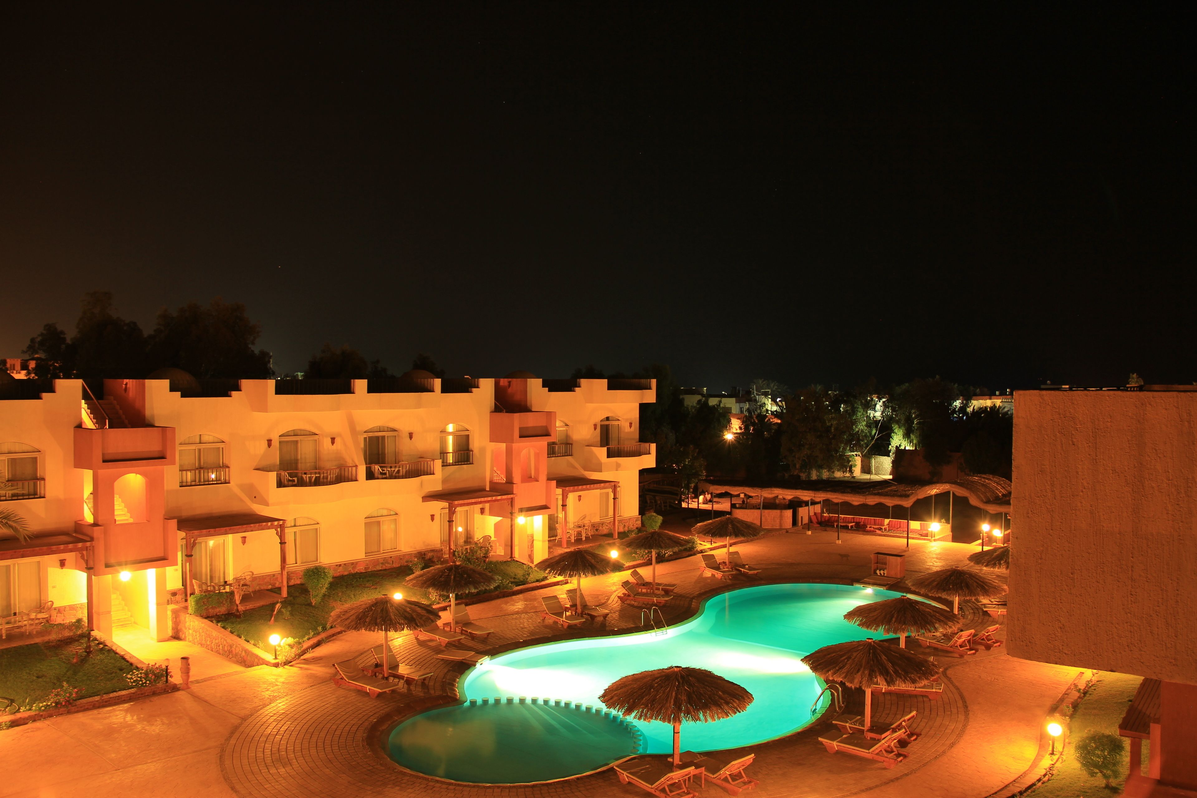 A nighttime view of a hotel with a swimming pool, lounge chairs, and umbrellas.