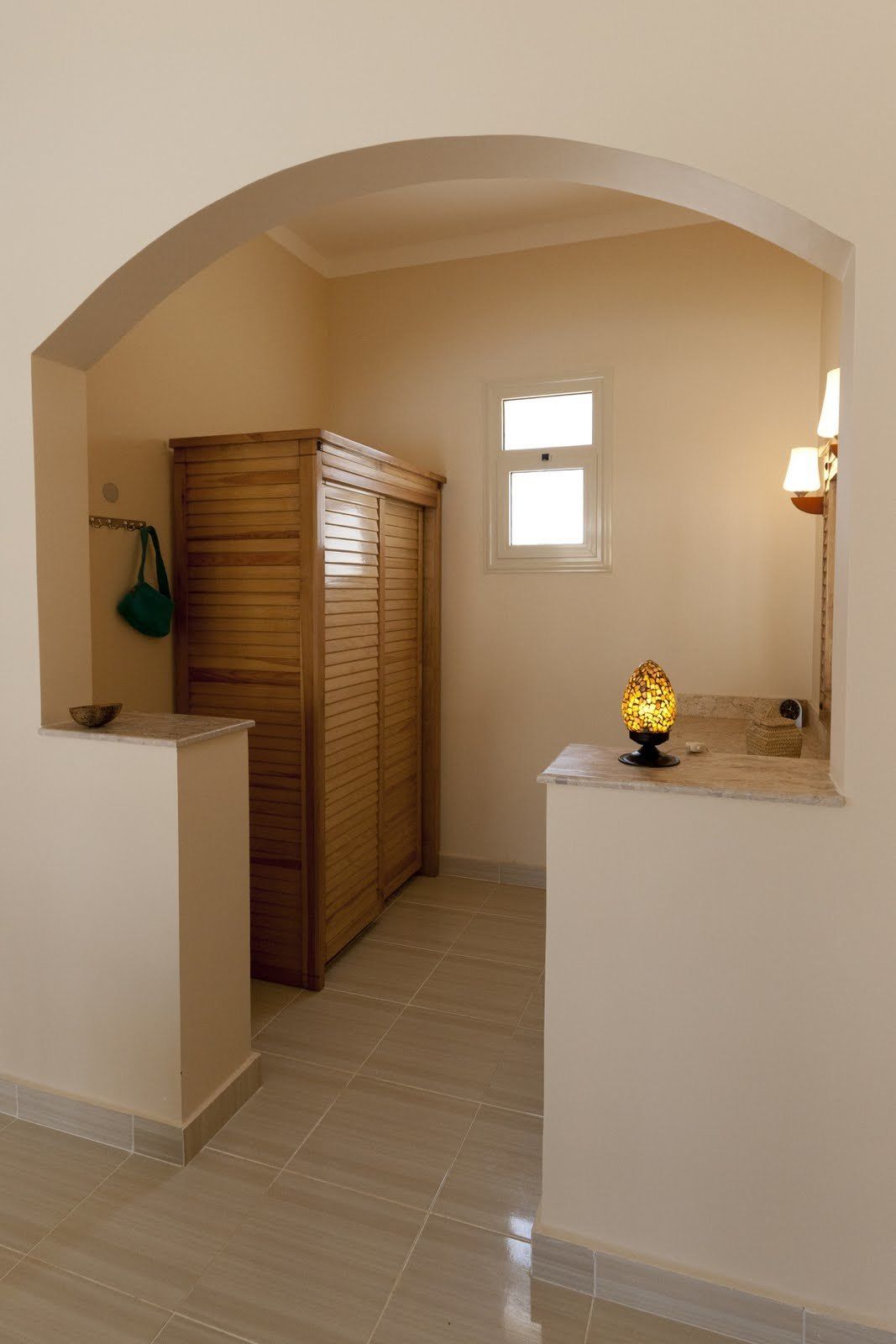 Una habitación con un armario de madera, una mesita con un cuenco, una lámpara y una ventana.
