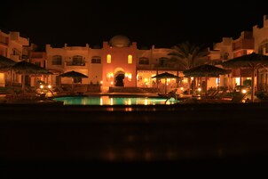 Vista nocturna de un complejo turístico con piscina, sillas de descanso, sombrillas y un edificio con cúpula y arcos.