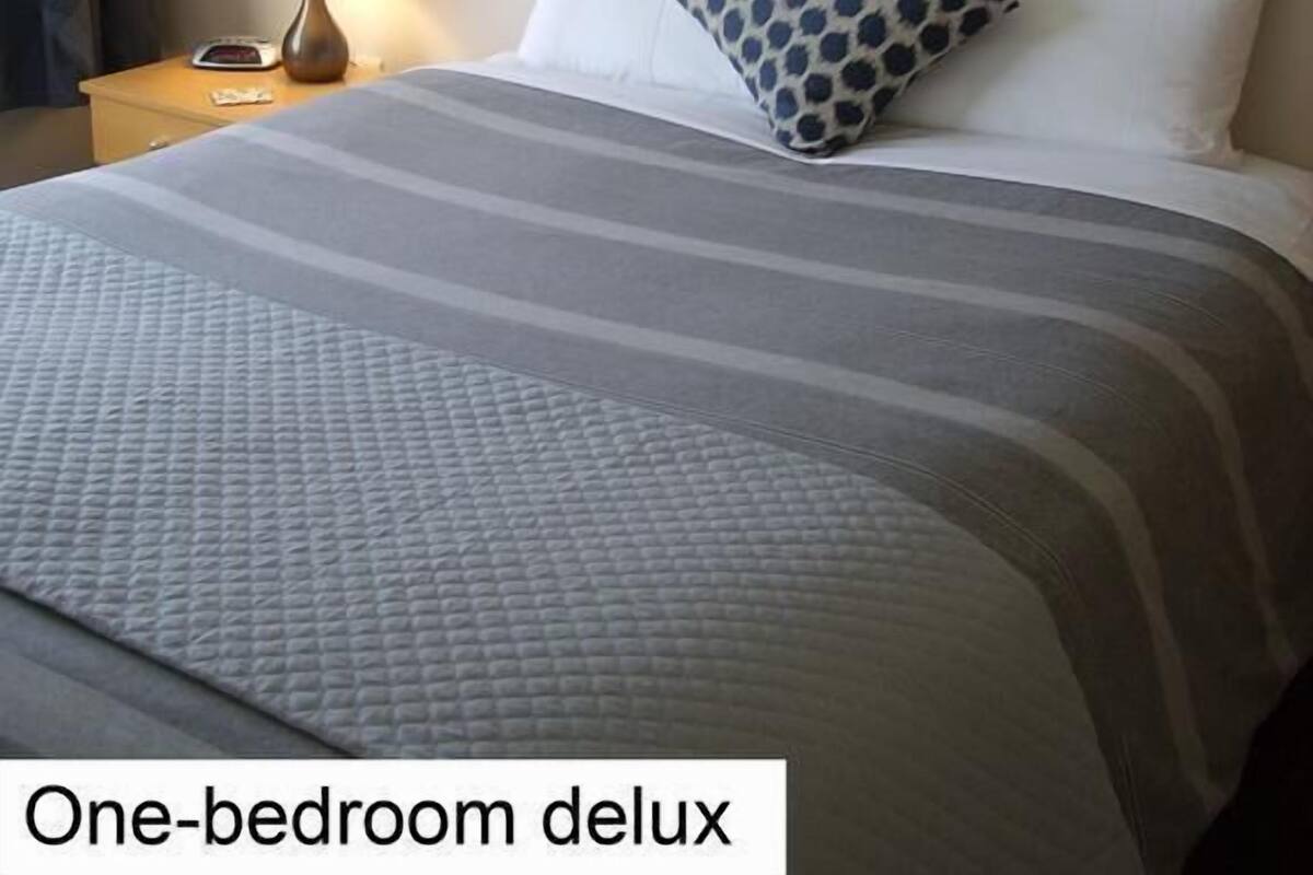 A one-bedroom deluxe room with a bed, pillows, a nightstand, a lamp, and a wall-mounted light fixture.