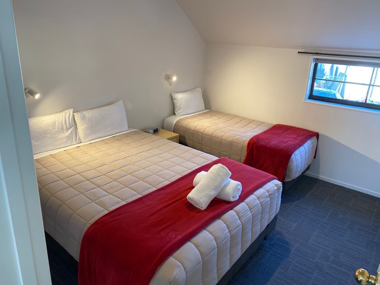Habitación de hotel con dos camas, cada una con una manta roja y almohadas blancas. Se ve una ventana con vistas al exterior.