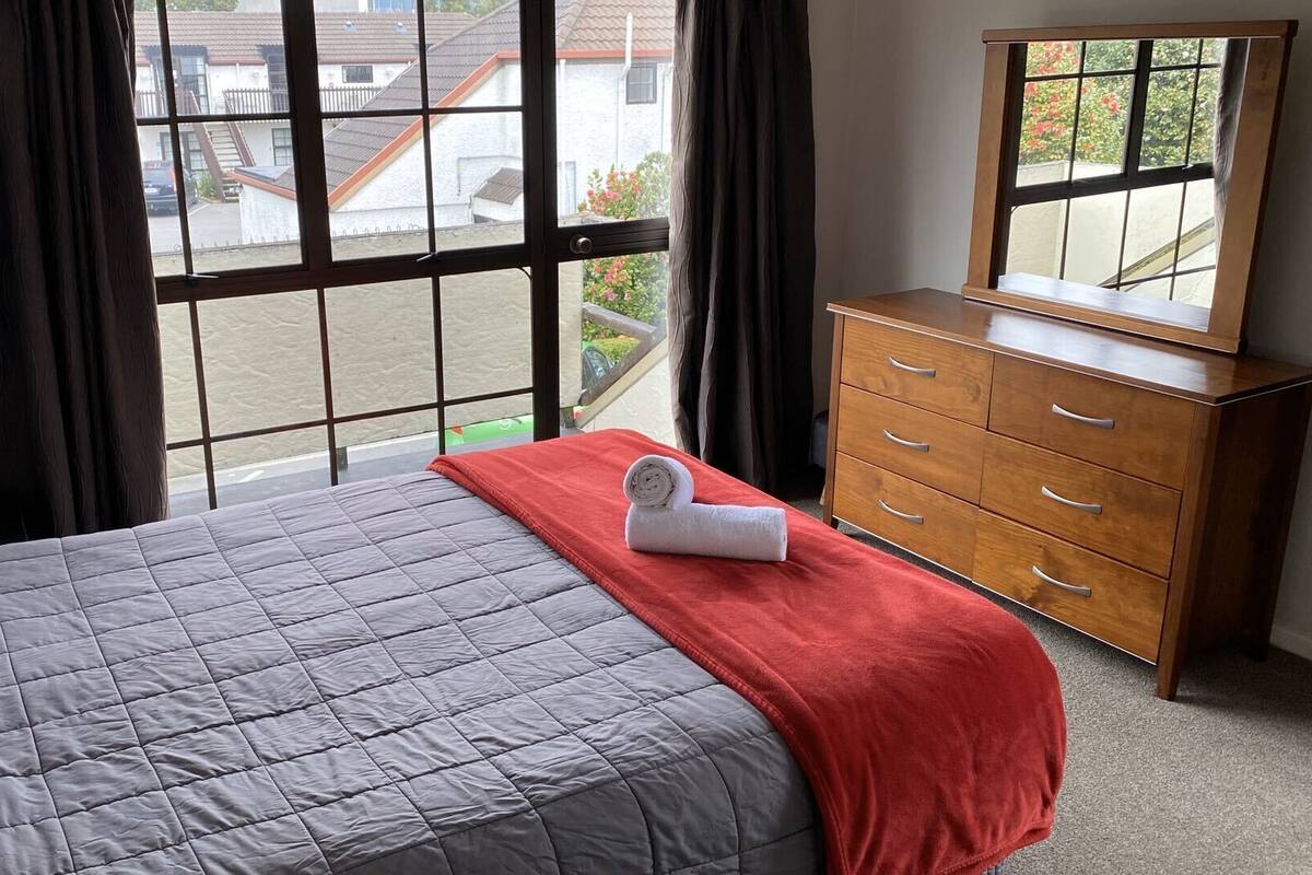 A hotel room with a bed, a wooden dresser, and a window with a view of buildings outside.