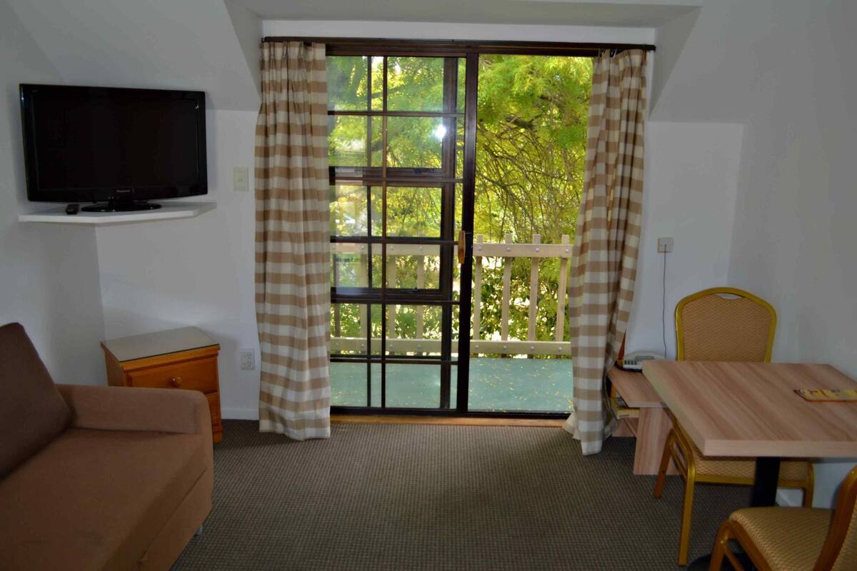 A room with a television on a shelf, a sofa, a table with chairs, and a window with curtains.