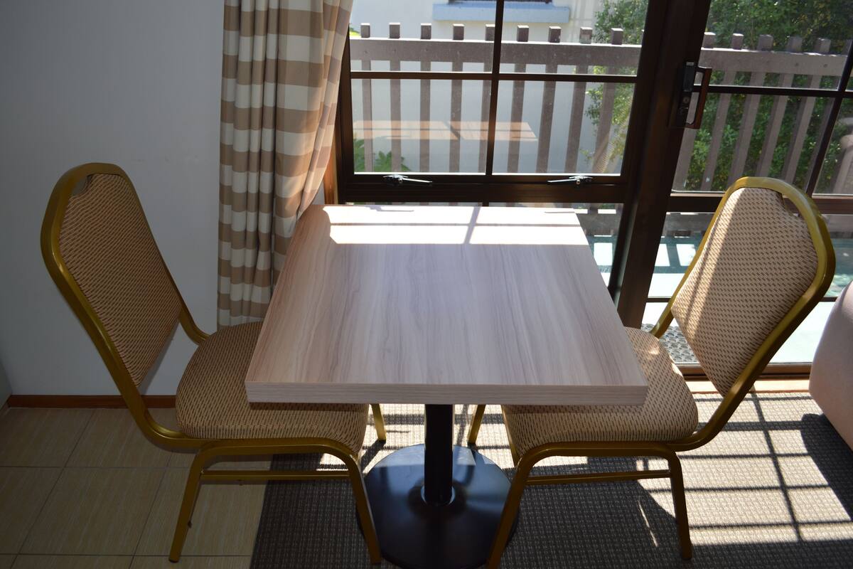 A dining area with a table and two chairs, a window with curtains, and a view of a wooden fence.