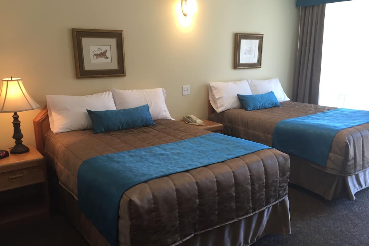 A hotel room with two beds, each with a blue bedspread and white pillows. There is a lamp on the left side, a nightstand with a phone, and framed pictures on the walls.