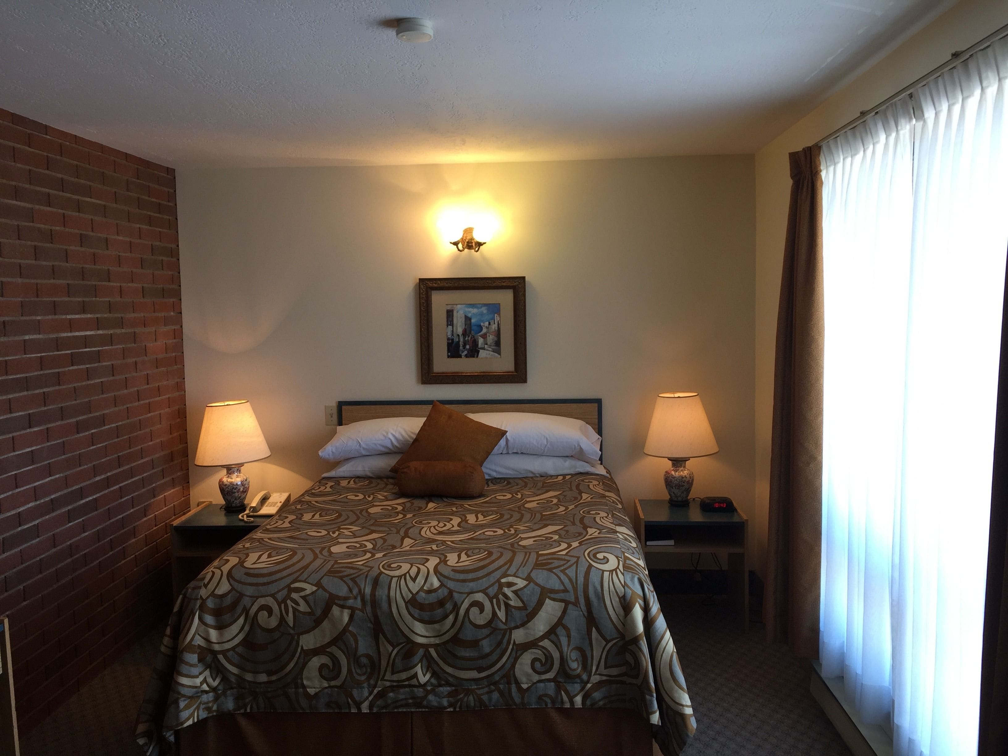 Une chambre d’hôtel avec un lit, deux lampes de chevet, une table de nuit, un tableau encadré au mur et une fenêtre avec des rideaux.
