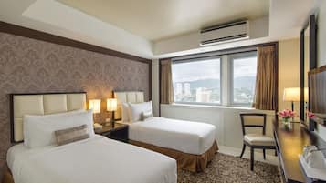 A hotel room with two beds, a desk with a chair, a window with a view of buildings, and an air conditioning unit.