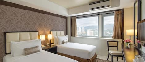A hotel room with two beds, a desk with a chair, a window with a view of buildings, and an air conditioning unit.