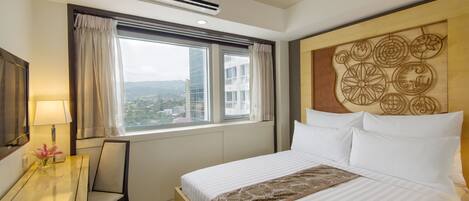 Una habitación de hotel con una cama grande, un escritorio con lámpara, una silla y vistas a edificios a través de la ventana.