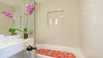 Un baño con una bañera llena de pétalos de rosa, una botella de champán en un balde y un espejo que refleja una orquídea púrpura.