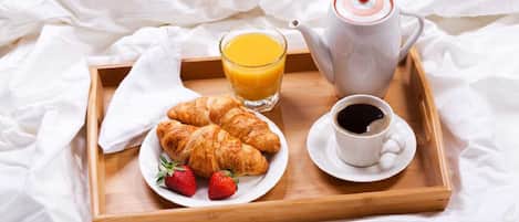 A wooden tray with croissants, strawberries, a glass of orange juice, a cup of coffee, and a teapot on a bed with white sheets.