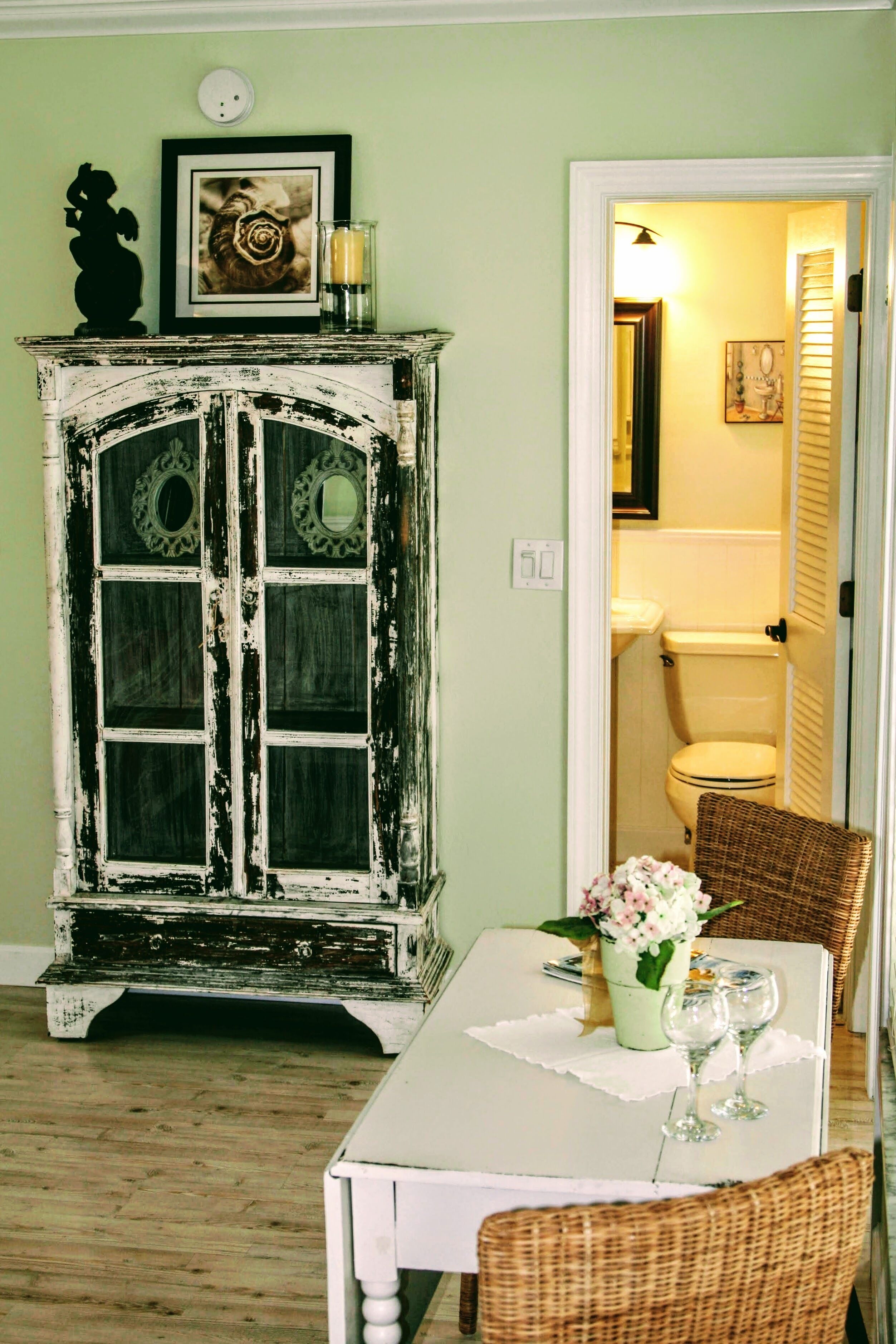 A dining area with a white table set for two, a wicker chair, a cabinet with decorative items, and a doorway leading to a bathroom.
