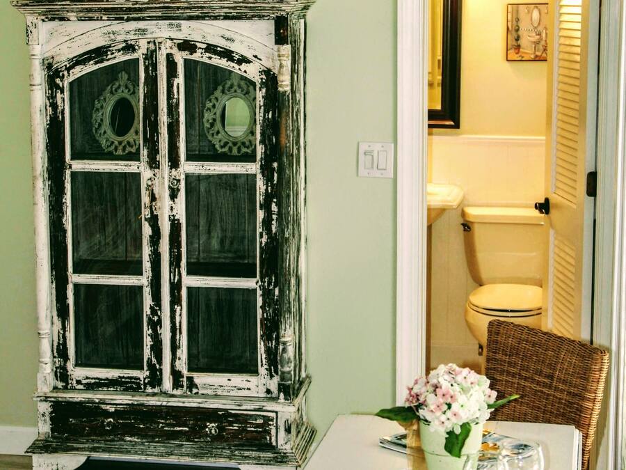 A dining area with a white table set for two, a wicker chair, a cabinet with decorative items, and a doorway leading to a bathroom.