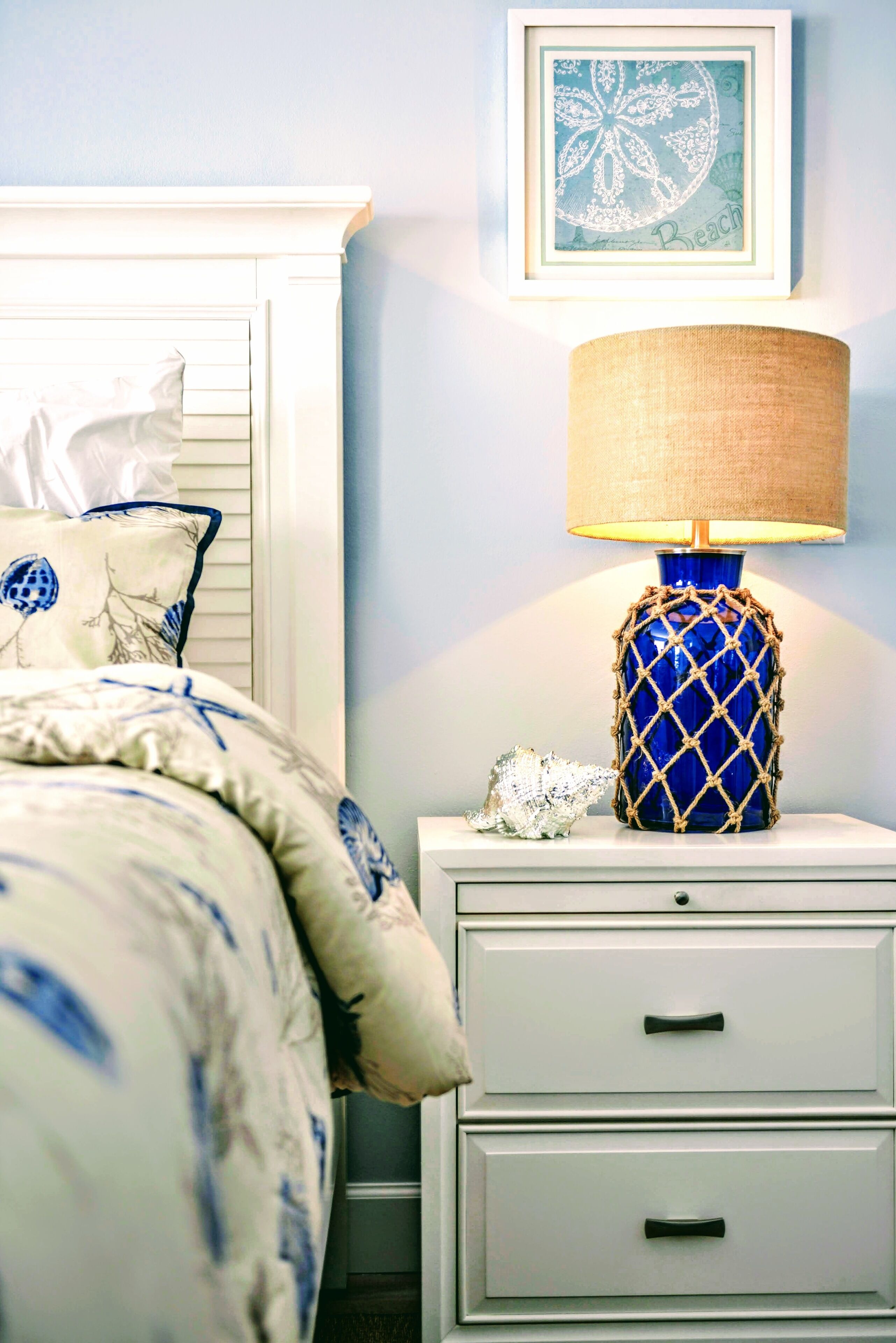 Uma cama bem arrumada com um edredom azul e branco estampado, uma mesa de cabeceira branca com um abajur azul e uma obra de arte emoldurada na parede.