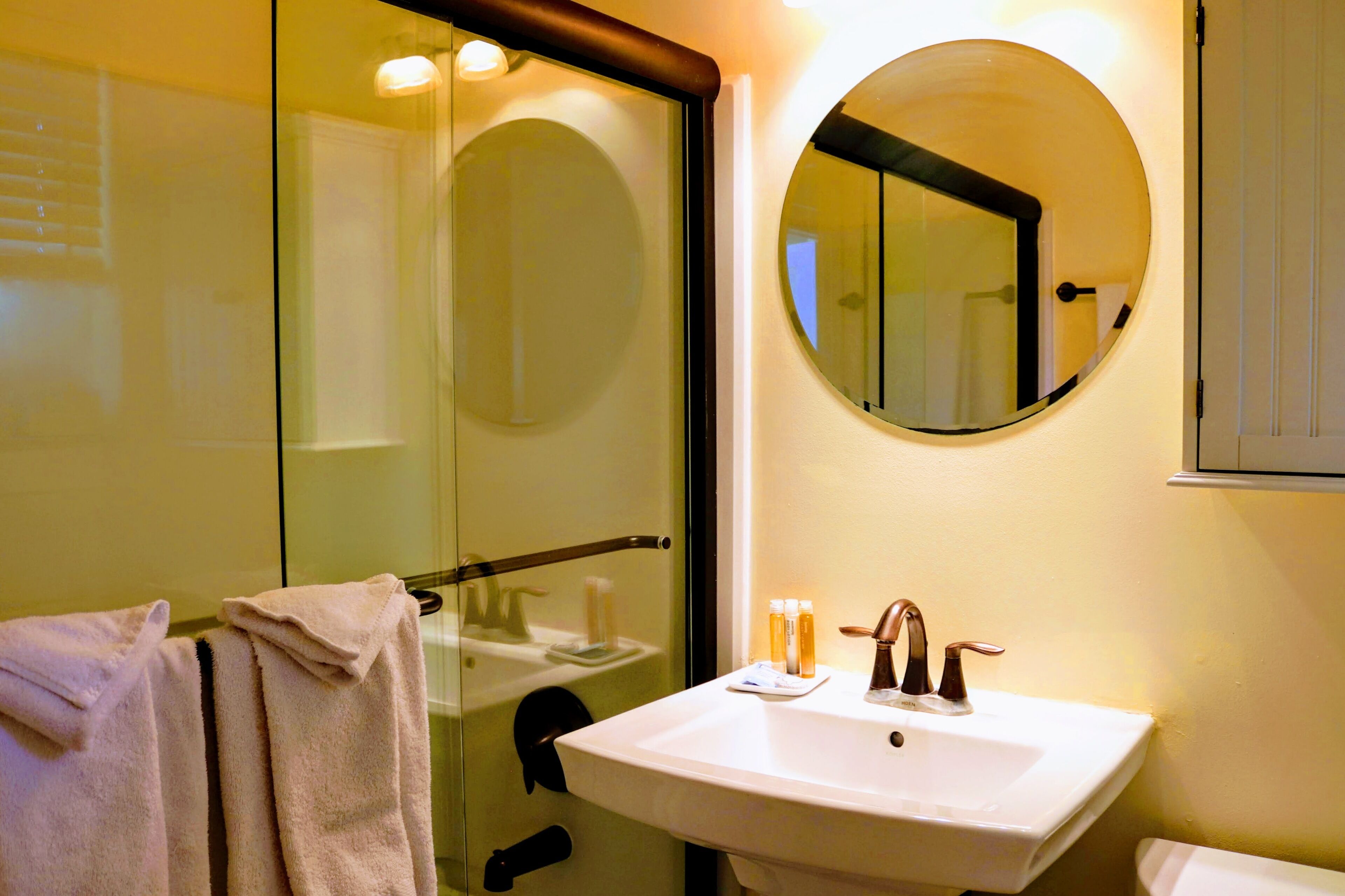 A bathroom with a glass shower door, a round mirror, a sink with a faucet, and a towel rack with towels.