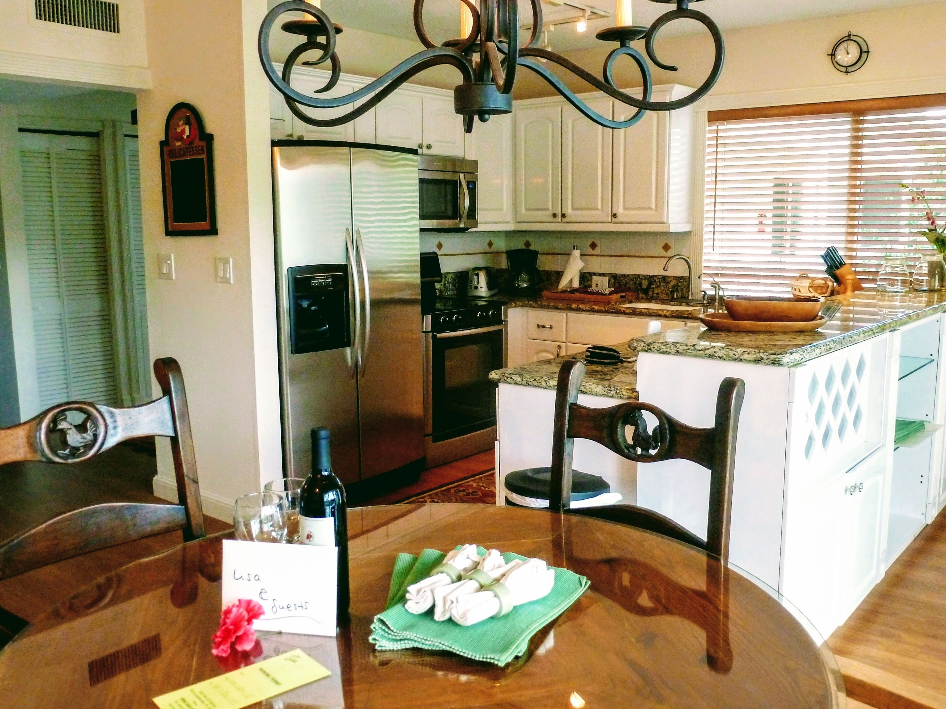 A dining area with a wooden table, chairs, a chandelier, and a kitchen in the background.