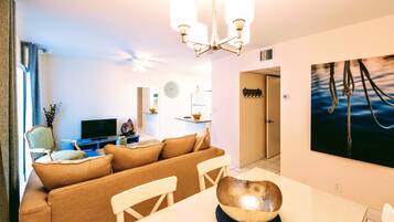 Ein modernes Wohnzimmer mit einer Couch, einem Esstisch und Stühlen. Es gibt einen Fernseher, einen Deckenventilator und Kunstwerke an den Wänden.