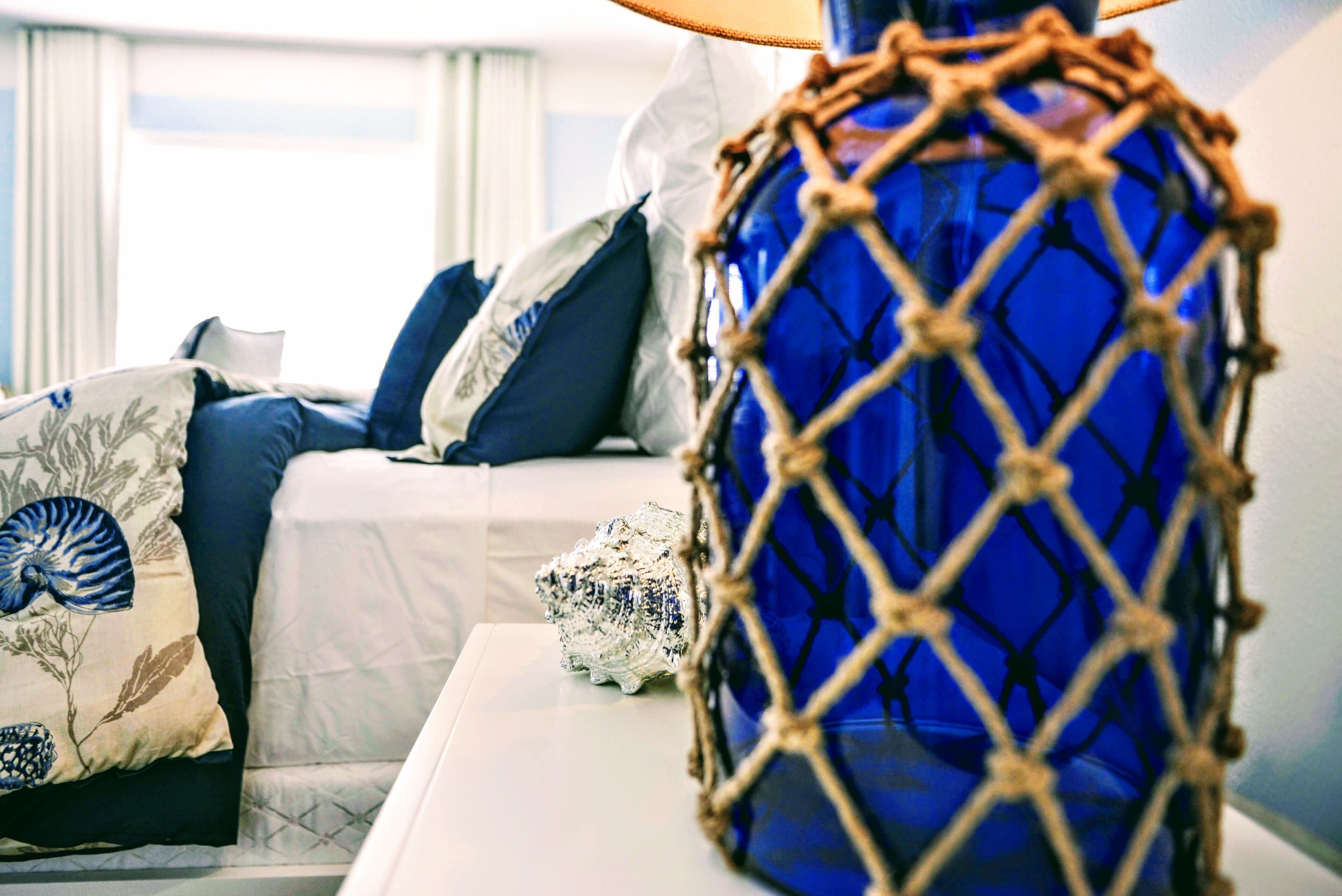 A bed with white bedding and blue pillows, a blue vase with a rope design, and a seashell on a bedside table.