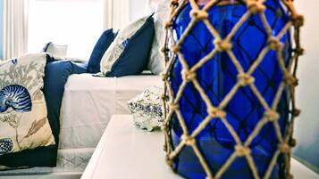 A bed with white bedding and blue pillows, a blue vase with a rope design, and a seashell on a bedside table.