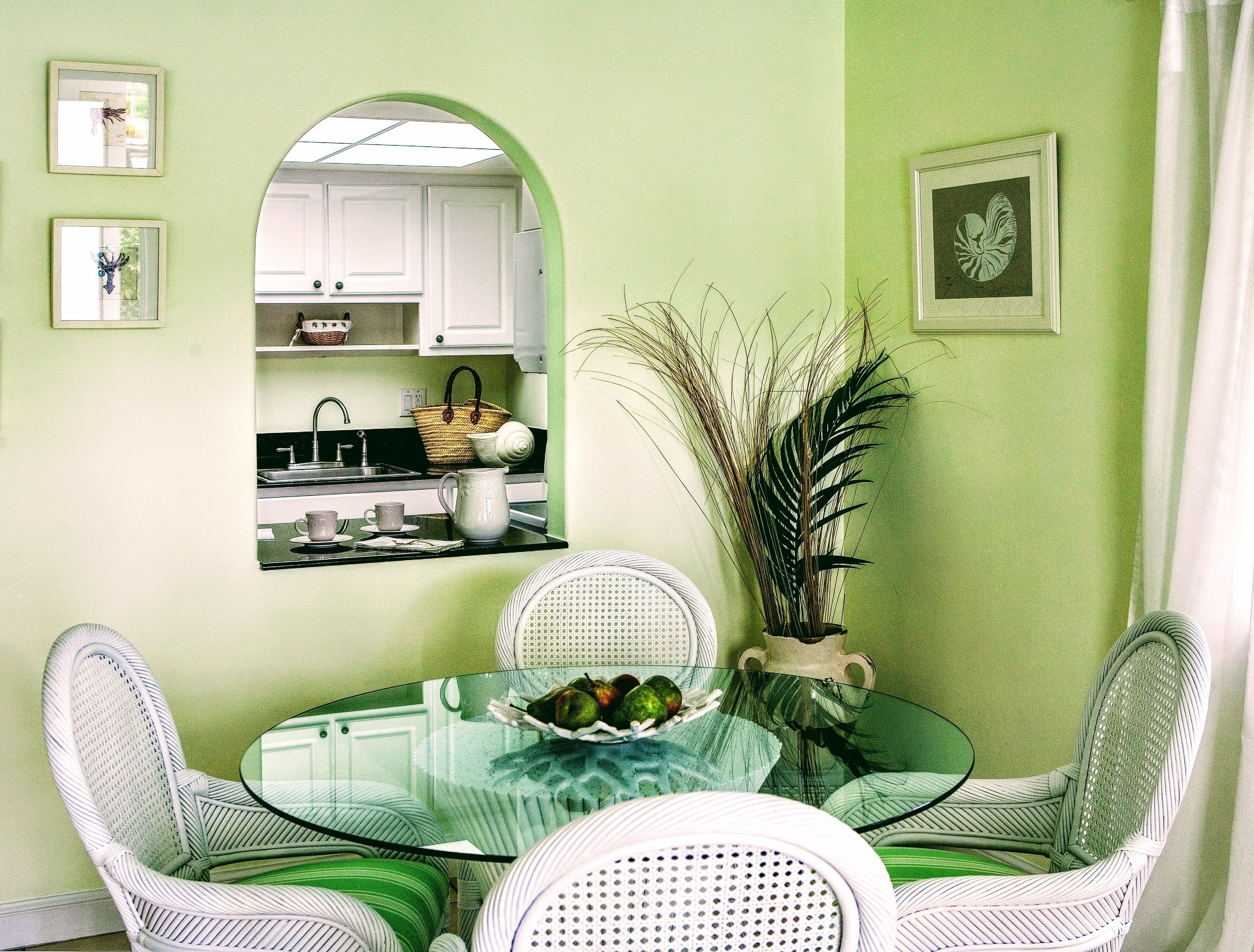 A dining area with a glass table surrounded by wicker chairs, a kitchen visible through an archway, and a potted plant on the table.