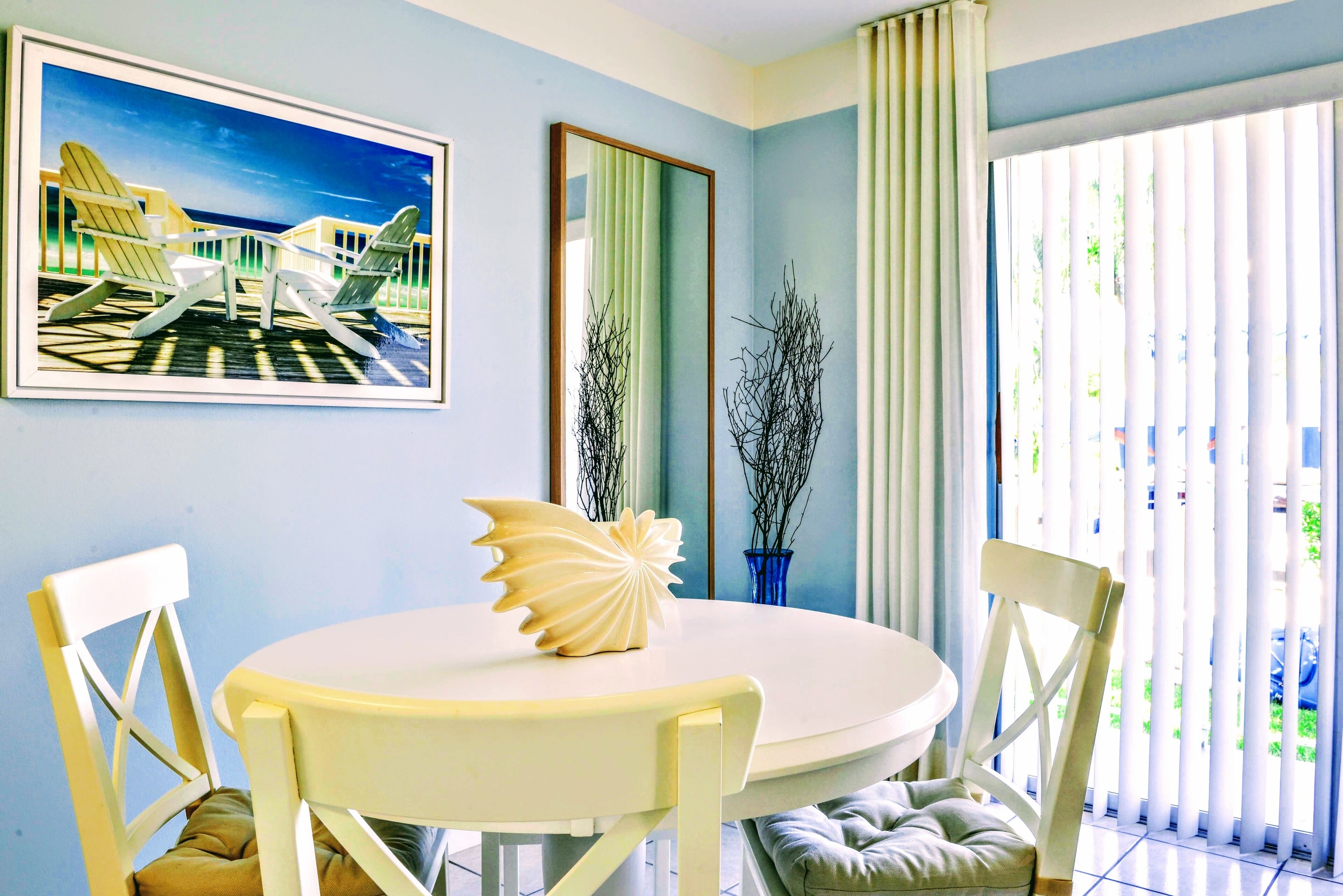 Een eethoek met een ronde tafel, witte stoelen, een decoratieve schelp als middelpunt en een schilderij van stoelen op een balkon.