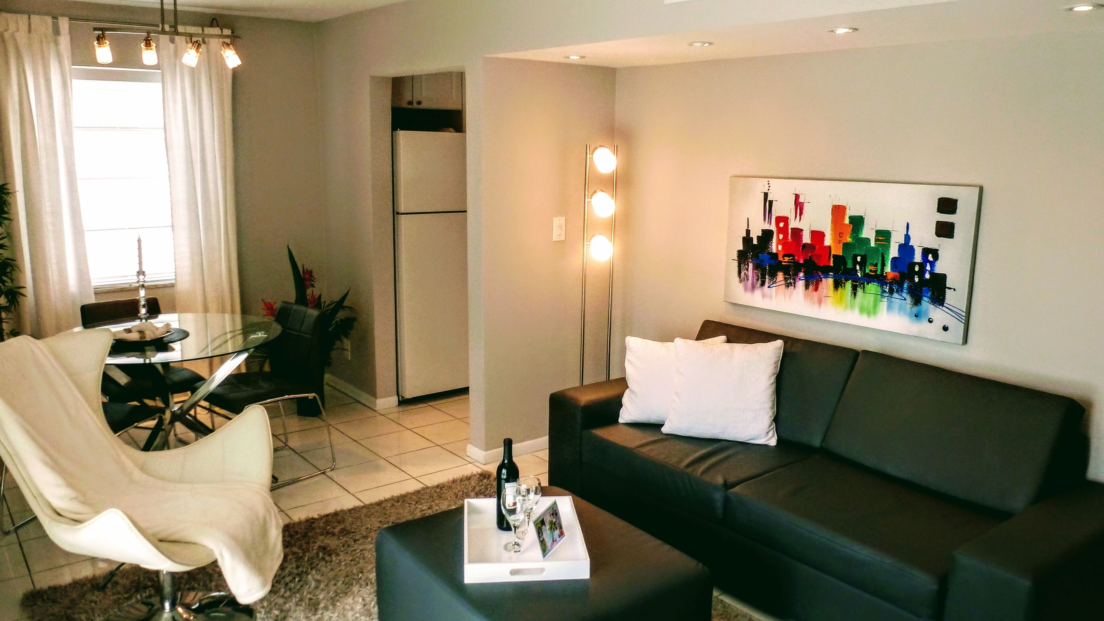 A modern living room with a dark sofa, a glass dining table, and a white chair. There is a painting on the wall and a bottle of wine on the coffee table.