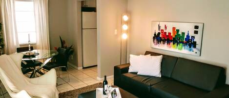 A modern living room with a dark sofa, a glass dining table, and a white chair. There is a painting on the wall and a bottle of wine on the coffee table.