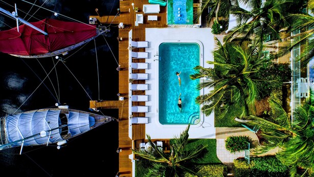 Vista aérea de un resort con piscina, sillas de descanso y dos veleros amarrados cerca.