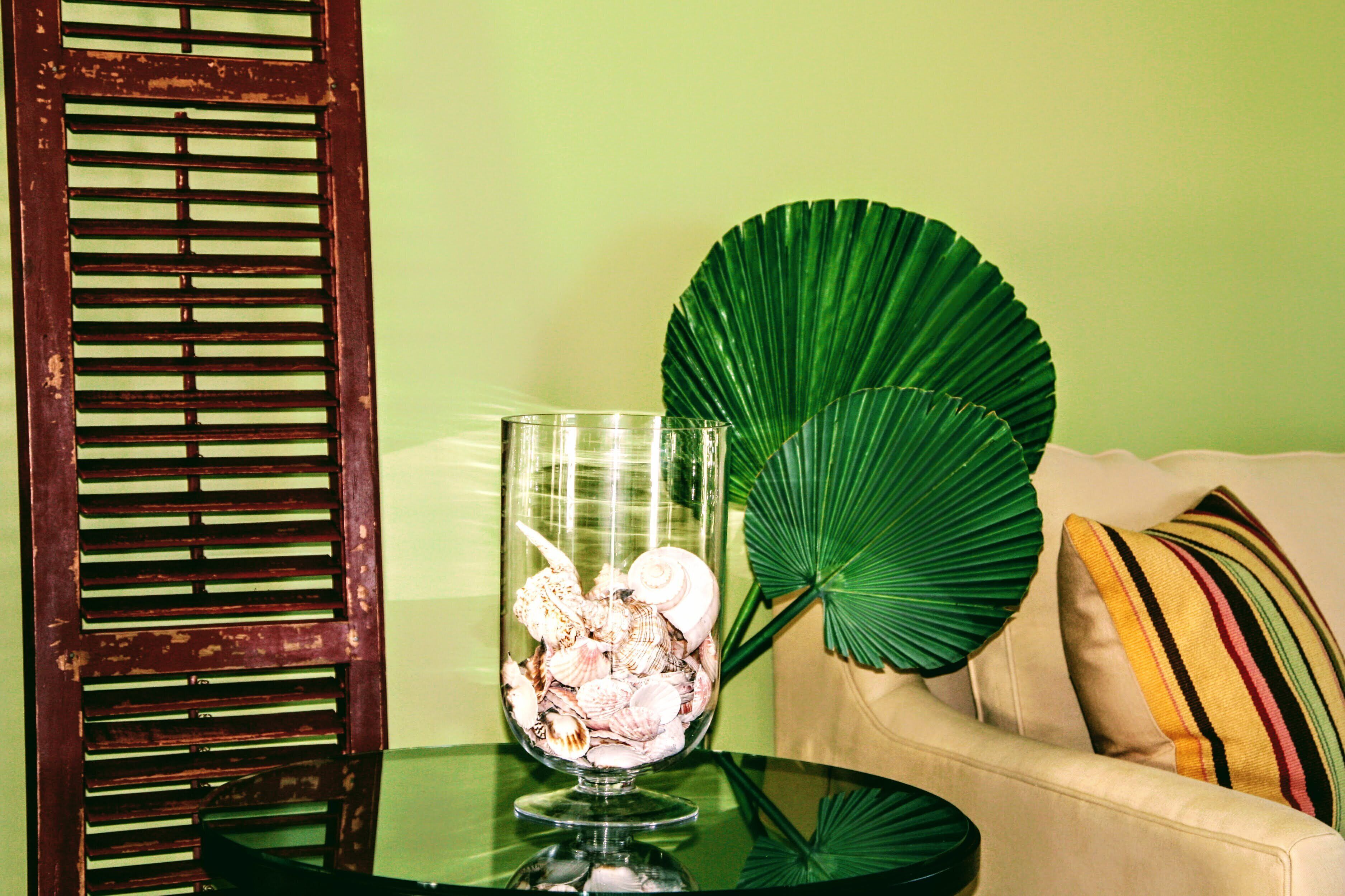 A glass bowl with seashells on a glass table, a large green leaf, and a beige sofa with striped pillows.