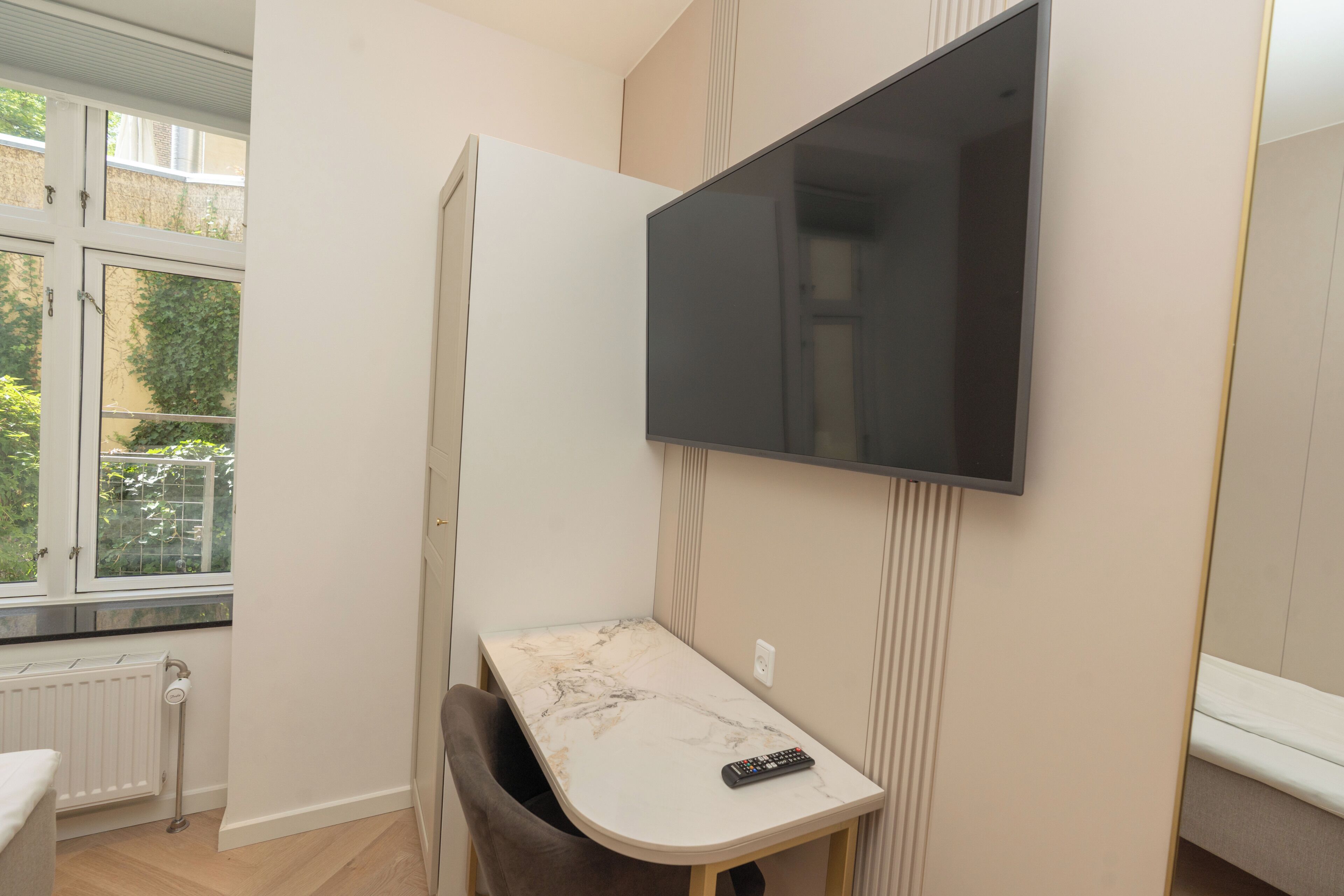 A small room with a TV mounted on the wall, a small table with a remote control, a radiator, and a window with a view of greenery.