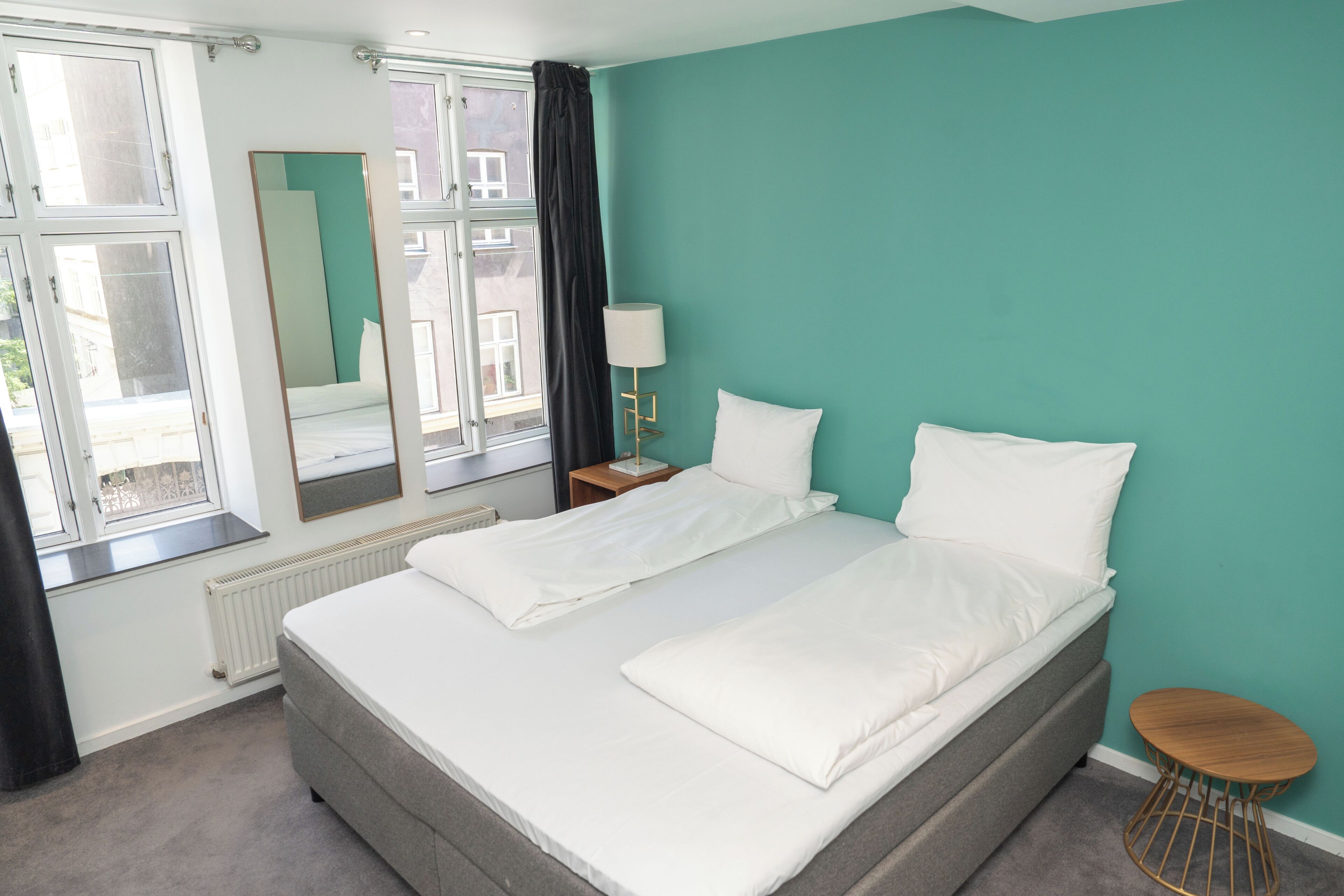 Een tweepersoonsbed met witte lakens, een nachtkastje met lamp, een ronde houten kruk en een raam met uitzicht op gebouwen.