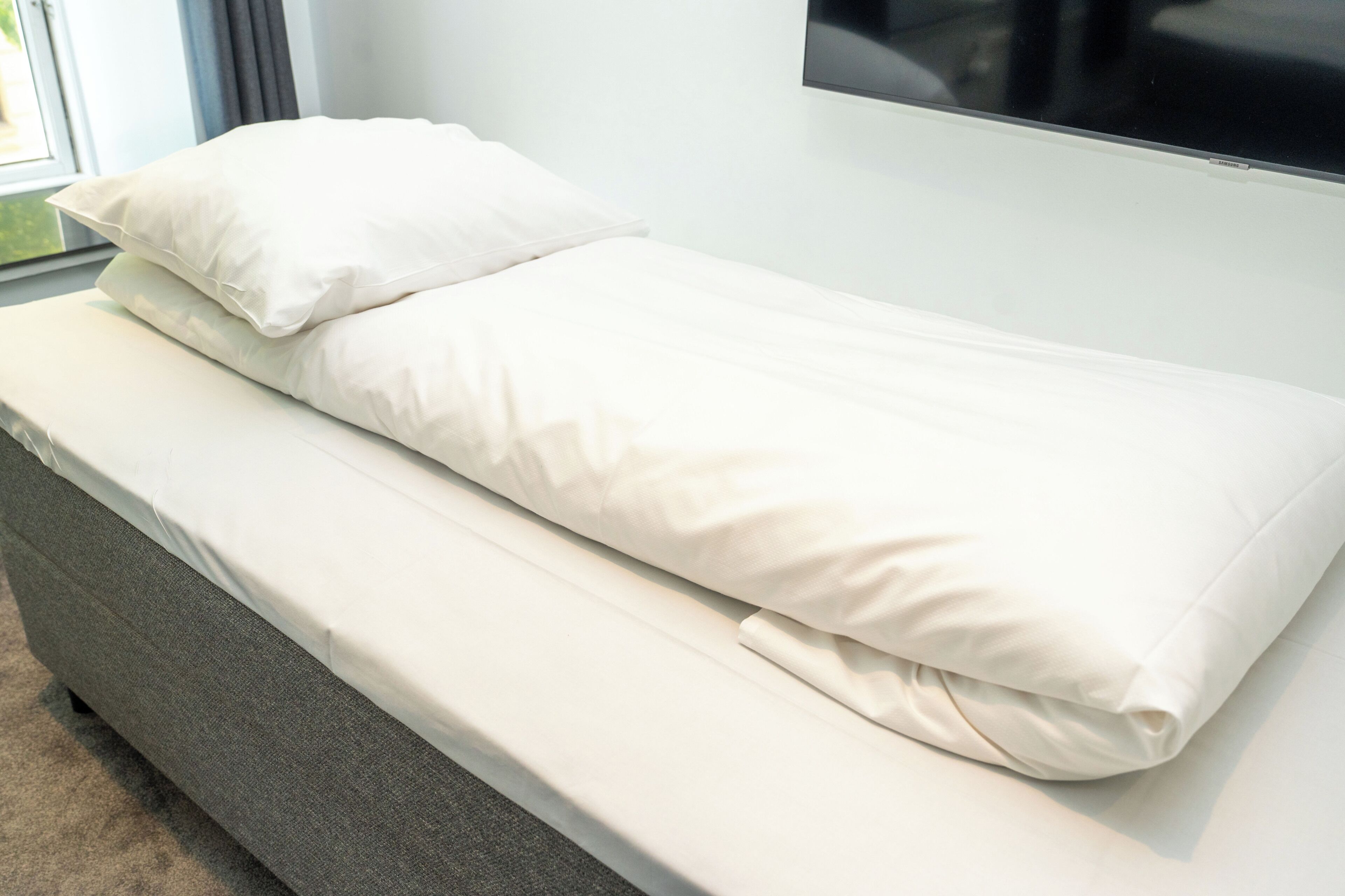 Ein Einzelbett mit weißer Bettwäsche, eine graue gepolsterte Sitzbank und ein großes Fenster mit Ausblick.