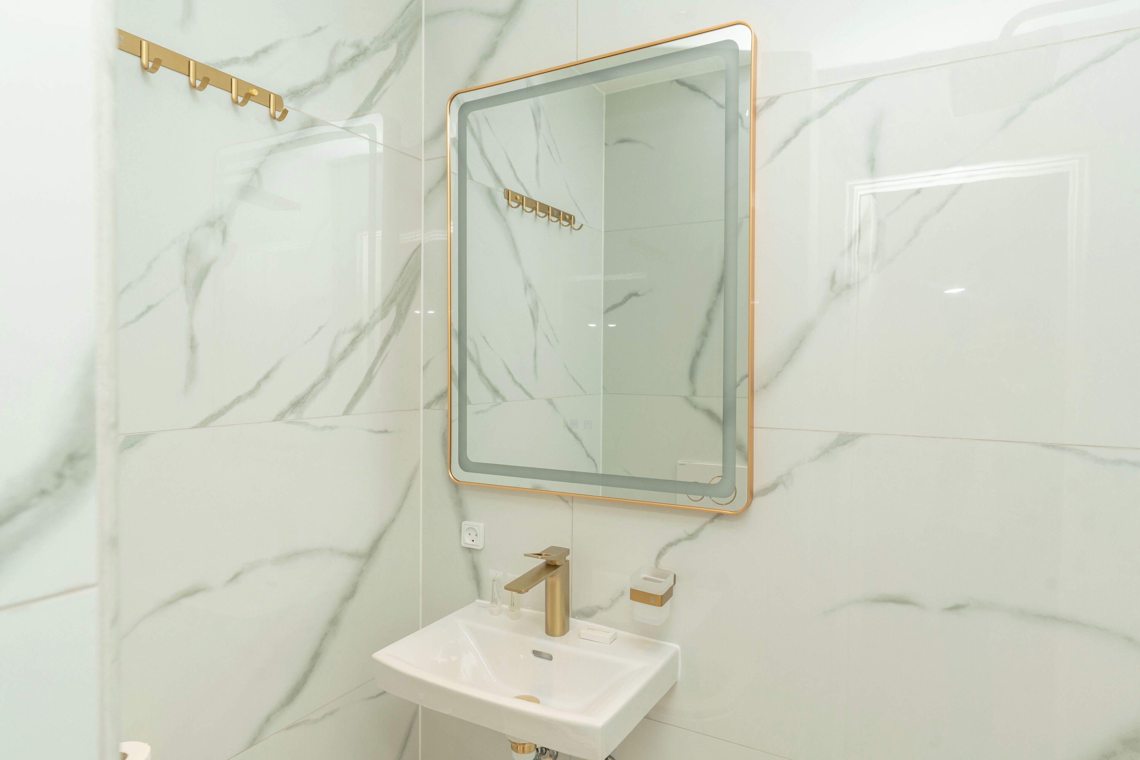 A bathroom with a marble wall, a gold-framed mirror, and a white sink with a gold faucet.