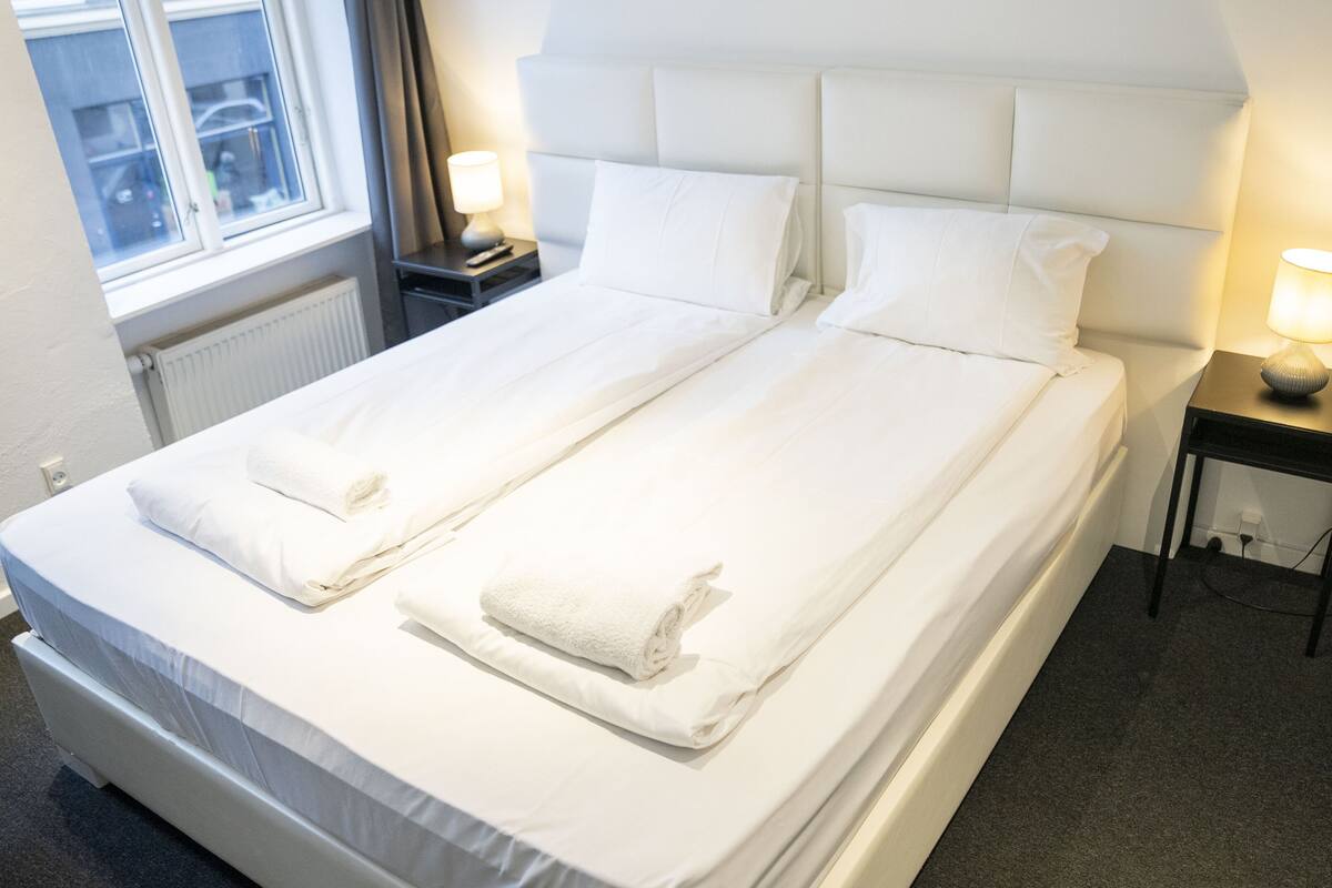 Una cama doble con sábanas blancas, dos mesitas de noche con lámparas, y una ventana con vistas al exterior.