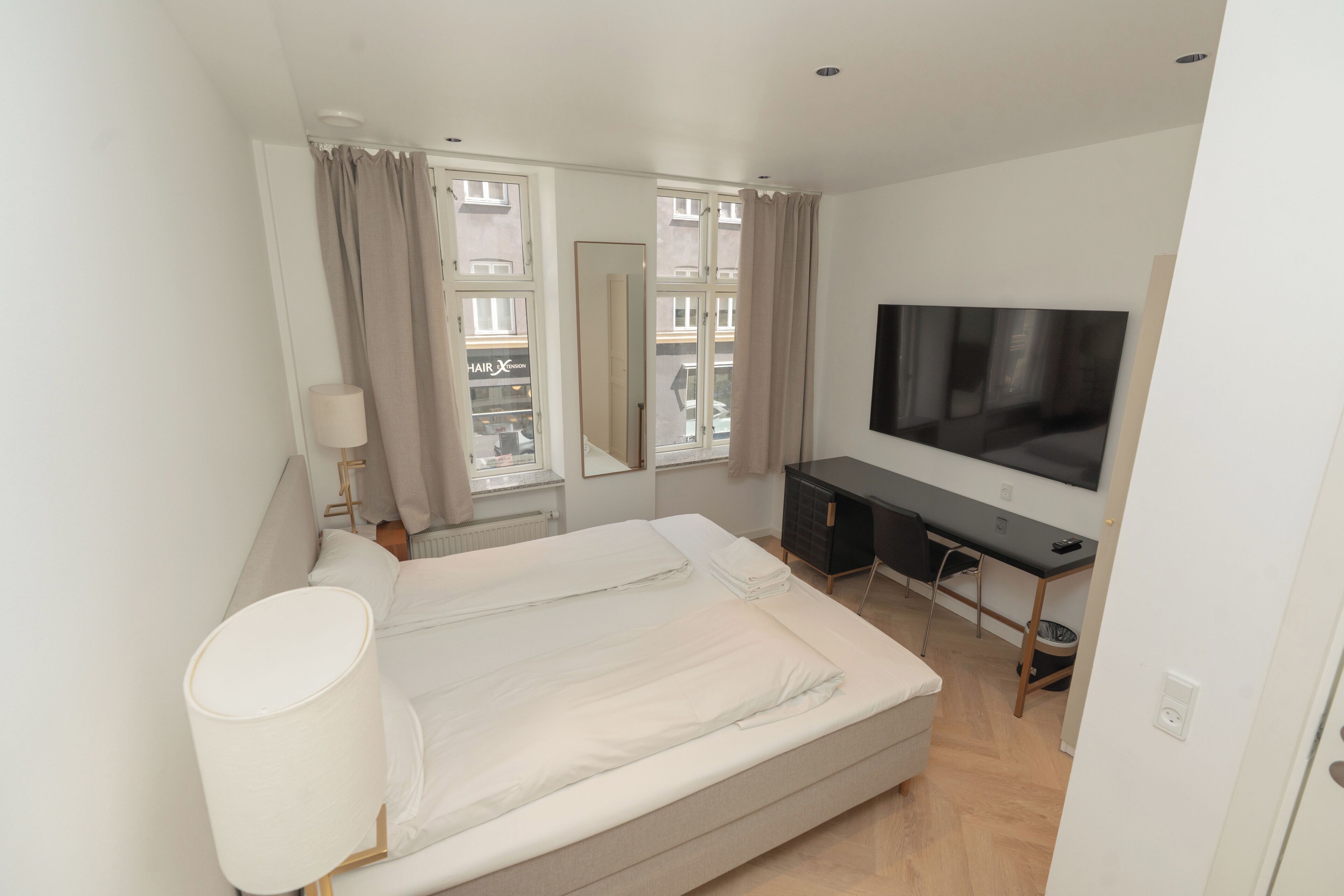 A bedroom with a bed, a TV, a desk, and a window with curtains.