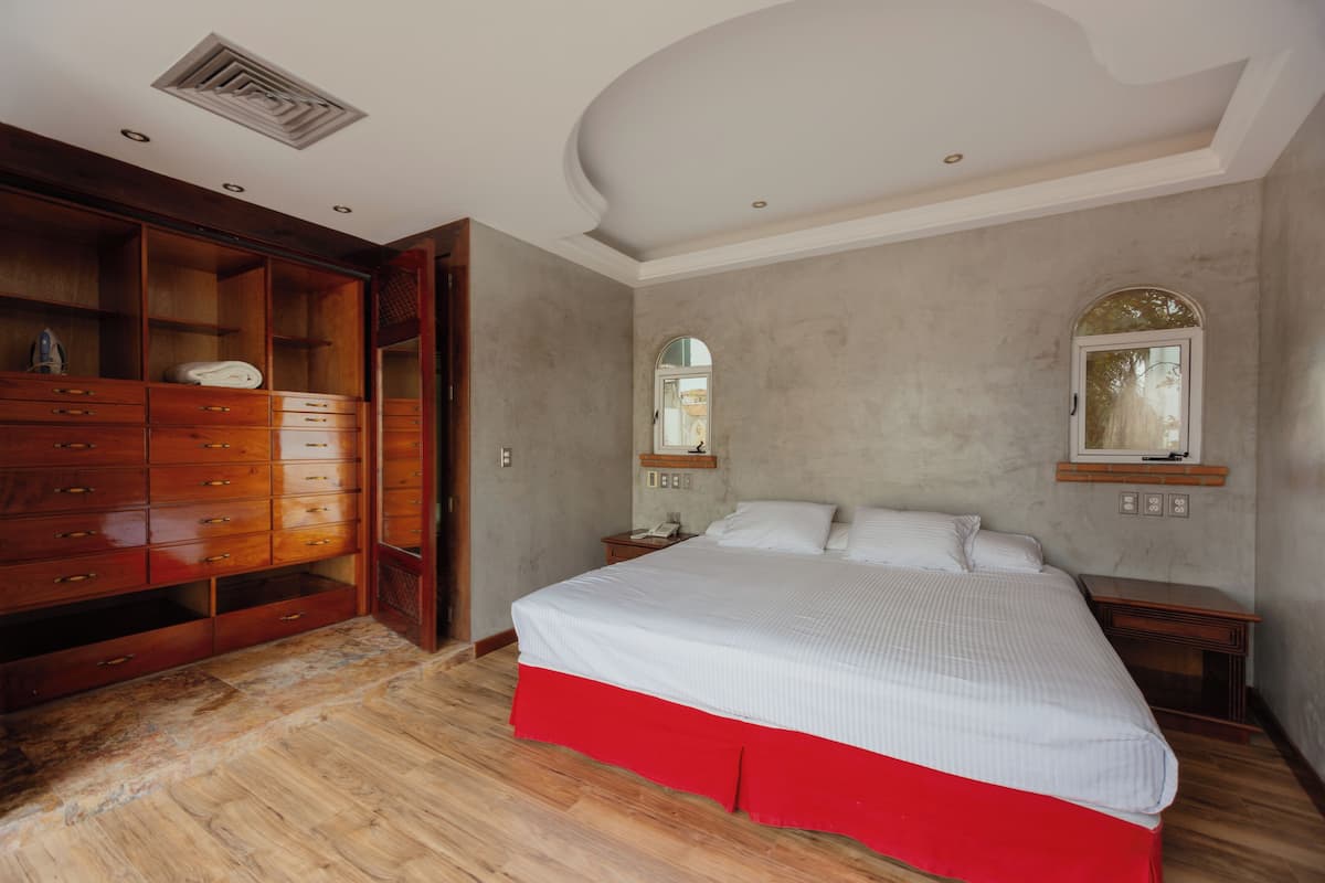 A bedroom with a large wooden wardrobe, a bed with a red skirt, and a small bedside table.