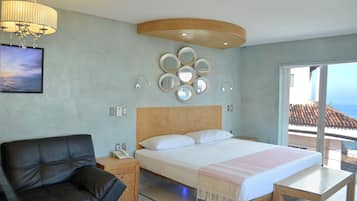 Un dormitorio con cama, cabecera de madera, mesita de noche, sillón de cuero negro, candelabro, espejos montados en la pared y vista al mar a través de una puerta corredera.