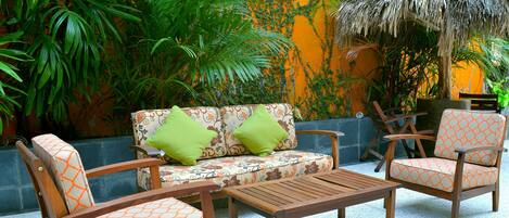 A patio area with wooden furniture, a patterned sofa, and lush greenery.