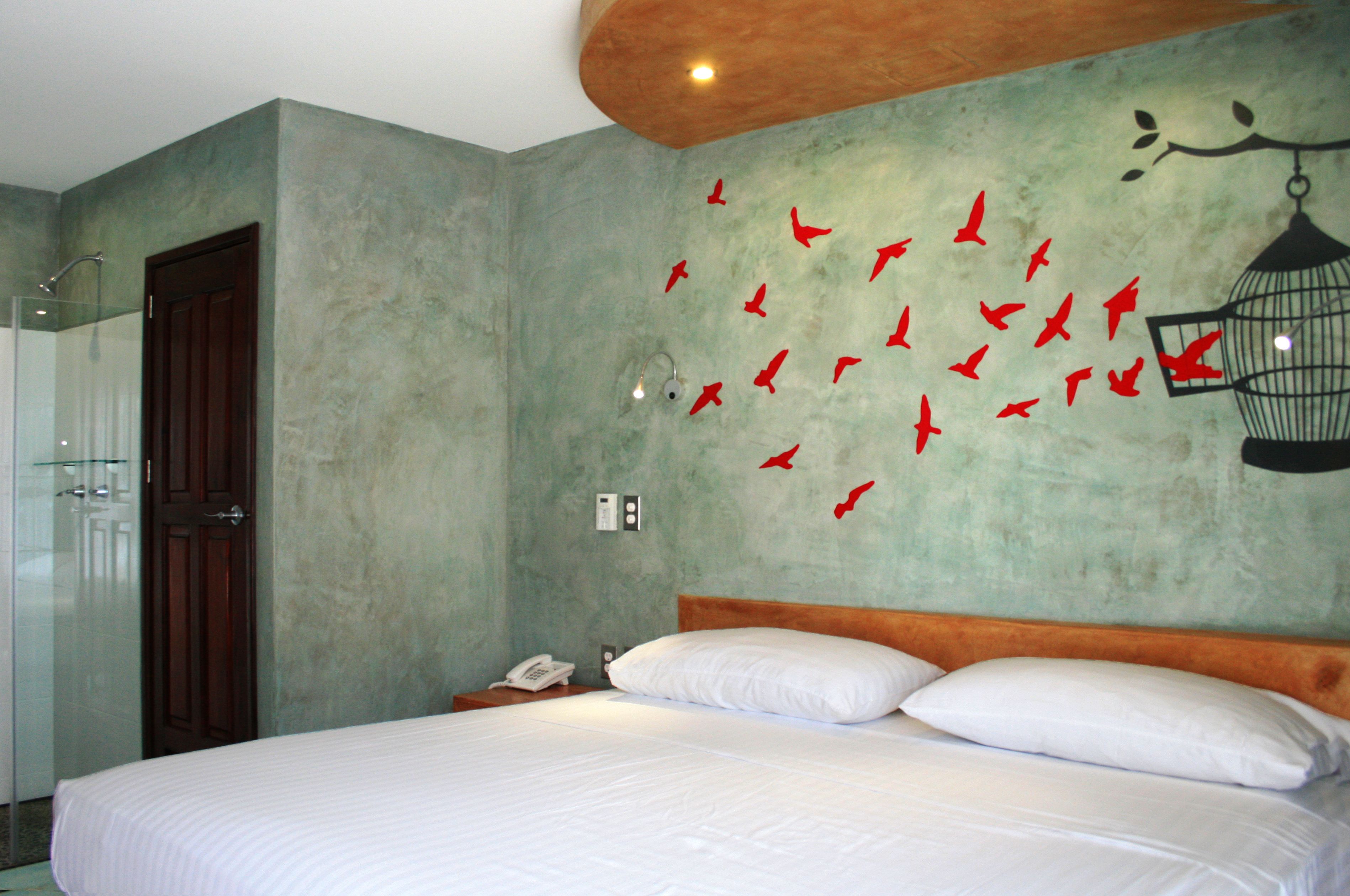 Habitación de hotel con una cama, cabecera de madera, mesita de noche con un teléfono y una pared decorada con diseños de pájaros rojos.