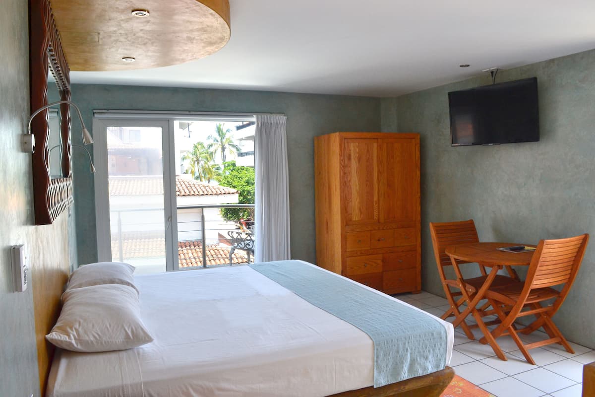 A bedroom with a bed, a wooden wardrobe, a TV mounted on the wall, and a balcony with a view of palm trees.