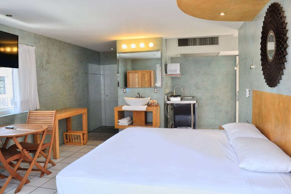 A hotel room with a bed, a desk with a sink, a mirror, and a window with curtains.