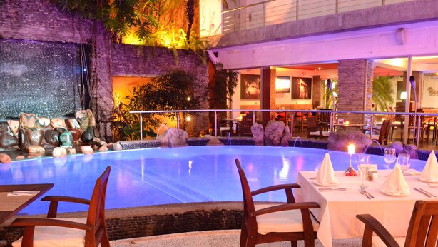A poolside dining area with tables set for two, surrounded by a stone wall and tropical plants. - Hotel Rio Malecon (Puerto Vallarta)