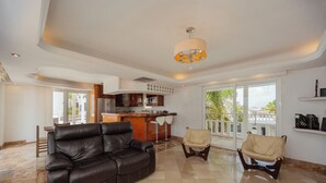 A spacious living area with a leather sofa, two armchairs, a kitchen in the background, and a large window offering a view of palm trees. - Hotel Rio Malecon (Puerto Vallarta)