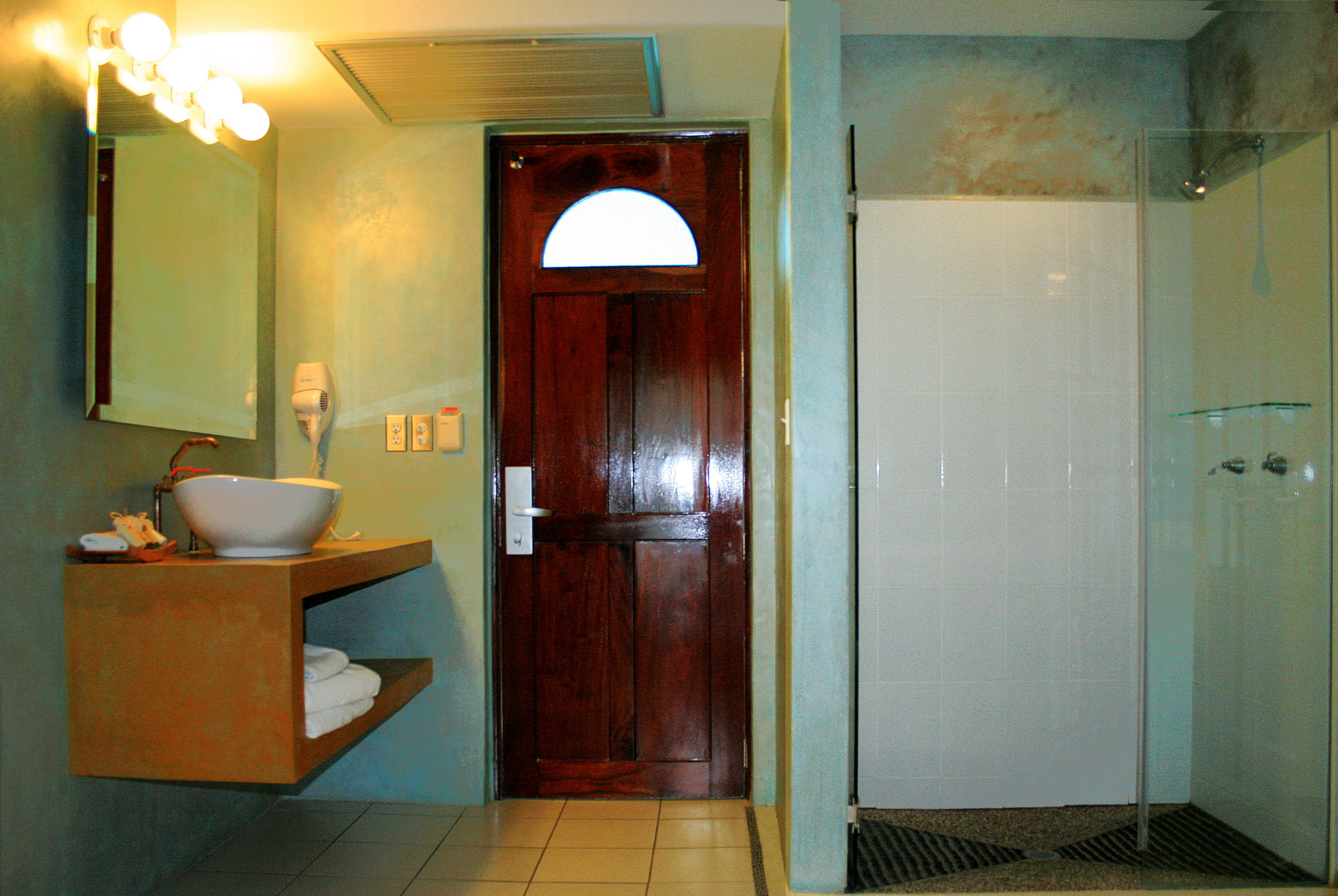 Un baño con puerta de madera, un lavabo con pileta, un espejo con luces y una zona de ducha.